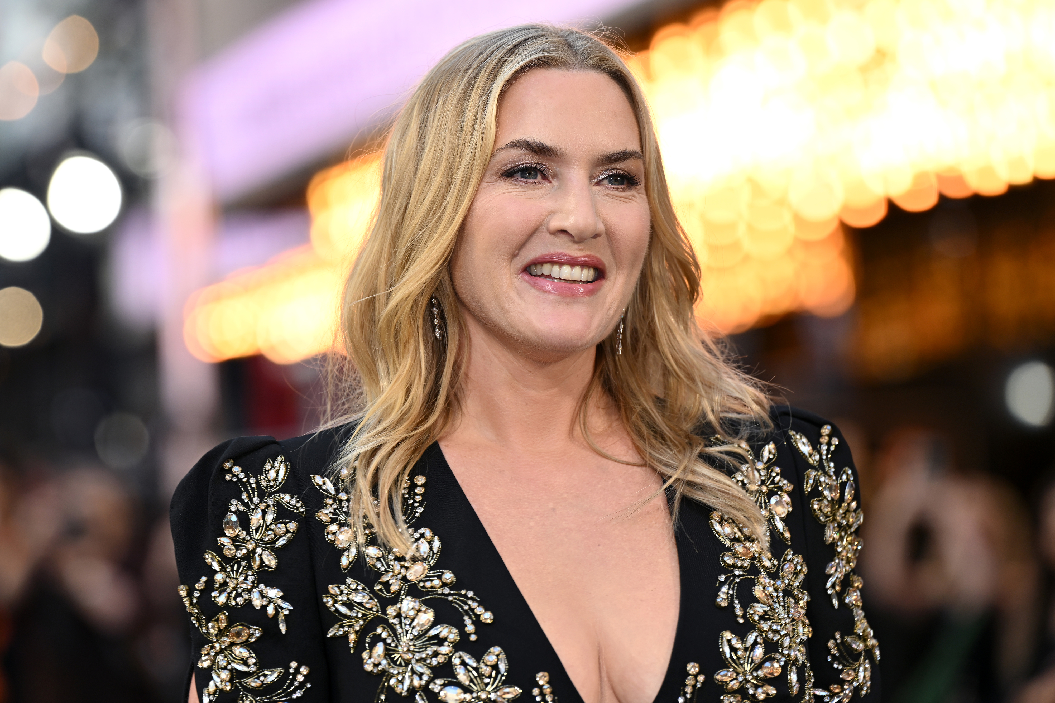 Kate Winslet smiles on the red carpet, wearing a dark dress adorned with intricate, gold floral embellishments