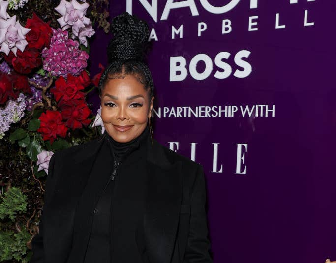 Janet Jackson at a Naomi Campbell event, wearing a dark outfit and standing in front of a floral display and a purple backdrop with event signage