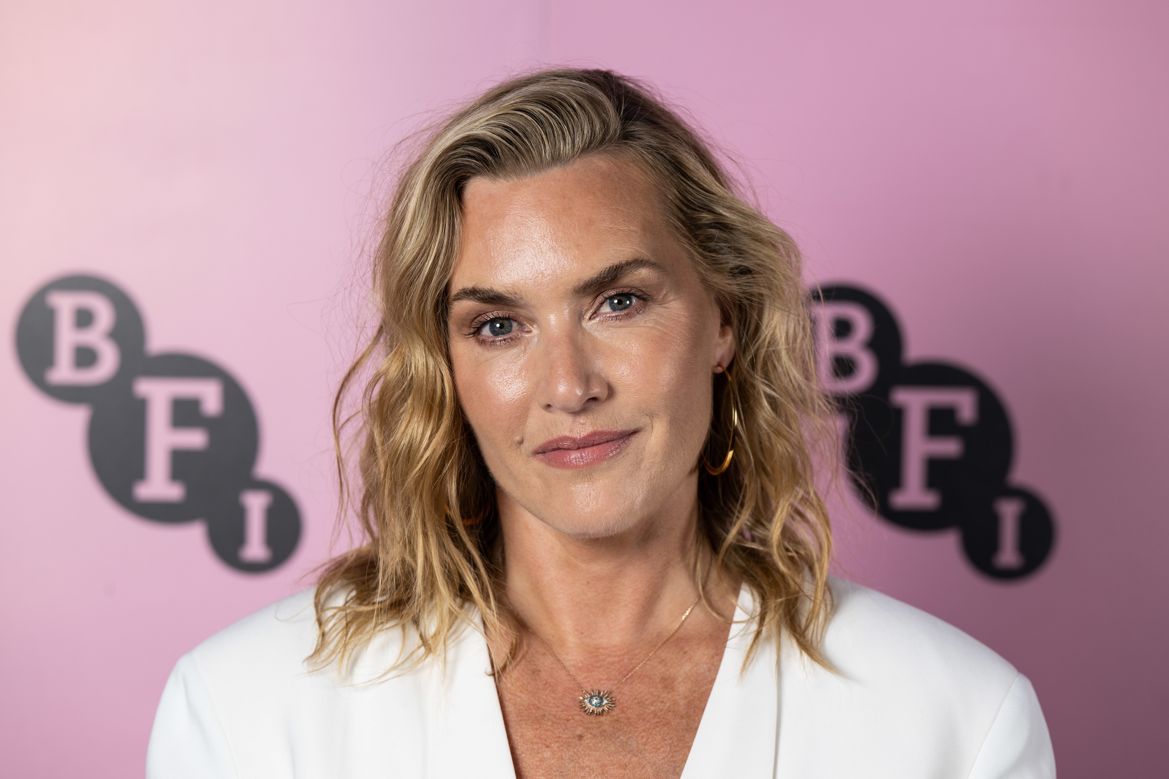 Kate Winslet at a BFI event, wearing a white blazer, gold hoop earrings, and a subtle necklace