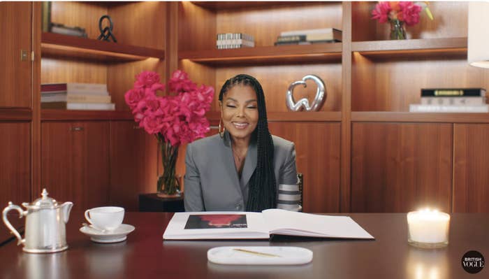 Janet Jackson, in a chic blazer, sits at a wooden desk with an open book, tea set, candle, and decorative bookshelves and flowers in the background