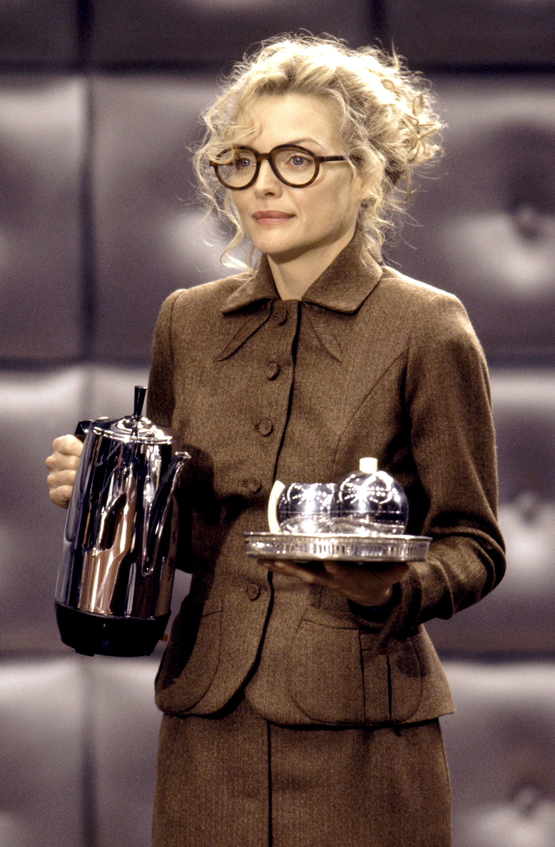 Michelle Pfeiffer as Selina Kyle in a suit and glasses, holding a coffee pot and a tray with cups from a scene in a movie