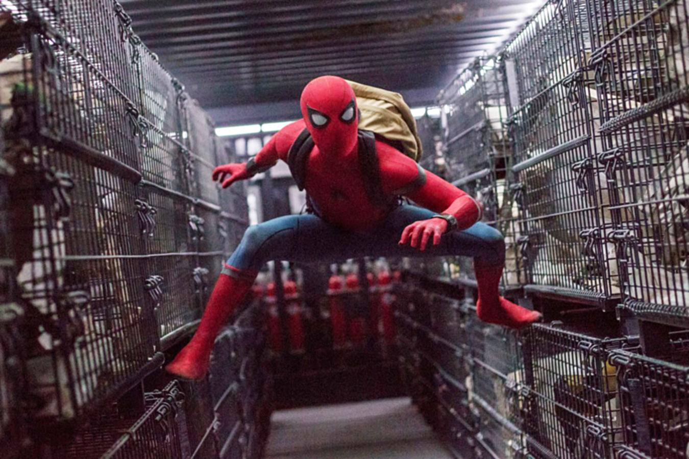 Spider-Man in costume is seen in an action pose, web-crawling inside a dimly lit, metallic, cage-filled space. He carries a backpack