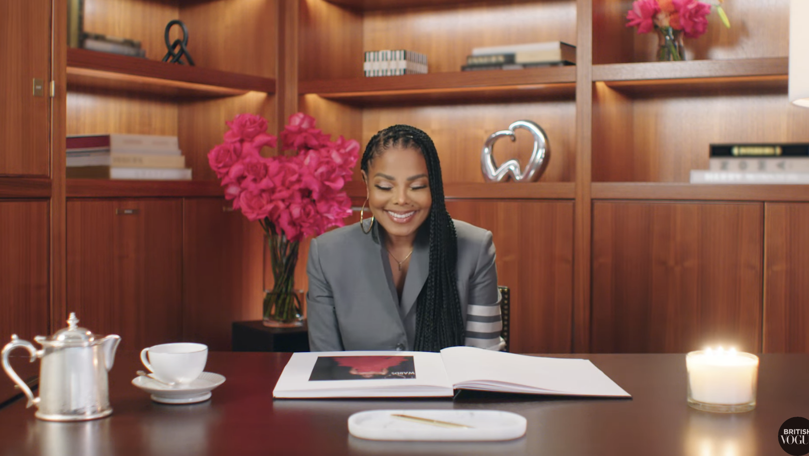 Janet Jackson sits at a desk with a book, smiling, surrounded by flowers and bookshelves in a warmly lit office
