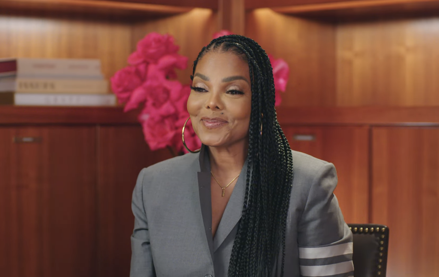 Janet Jackson in a grey blazer with striped sleeves, sitting in front of wooden shelves adorned with pink flowers