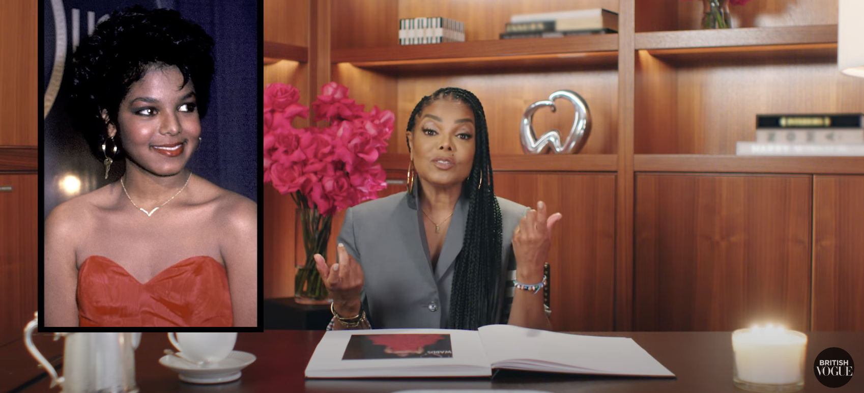Janet Jackson sits at a desk, next to a framed photo of her younger self in a strapless dress. Shelves with books and decor are visible in the background
