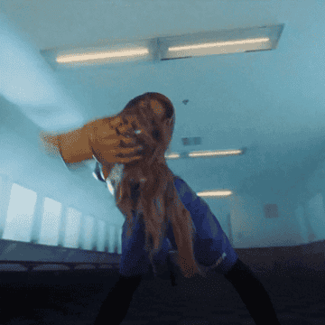 A person with long hair dances energetically in a room with fluorescent lights on the ceiling