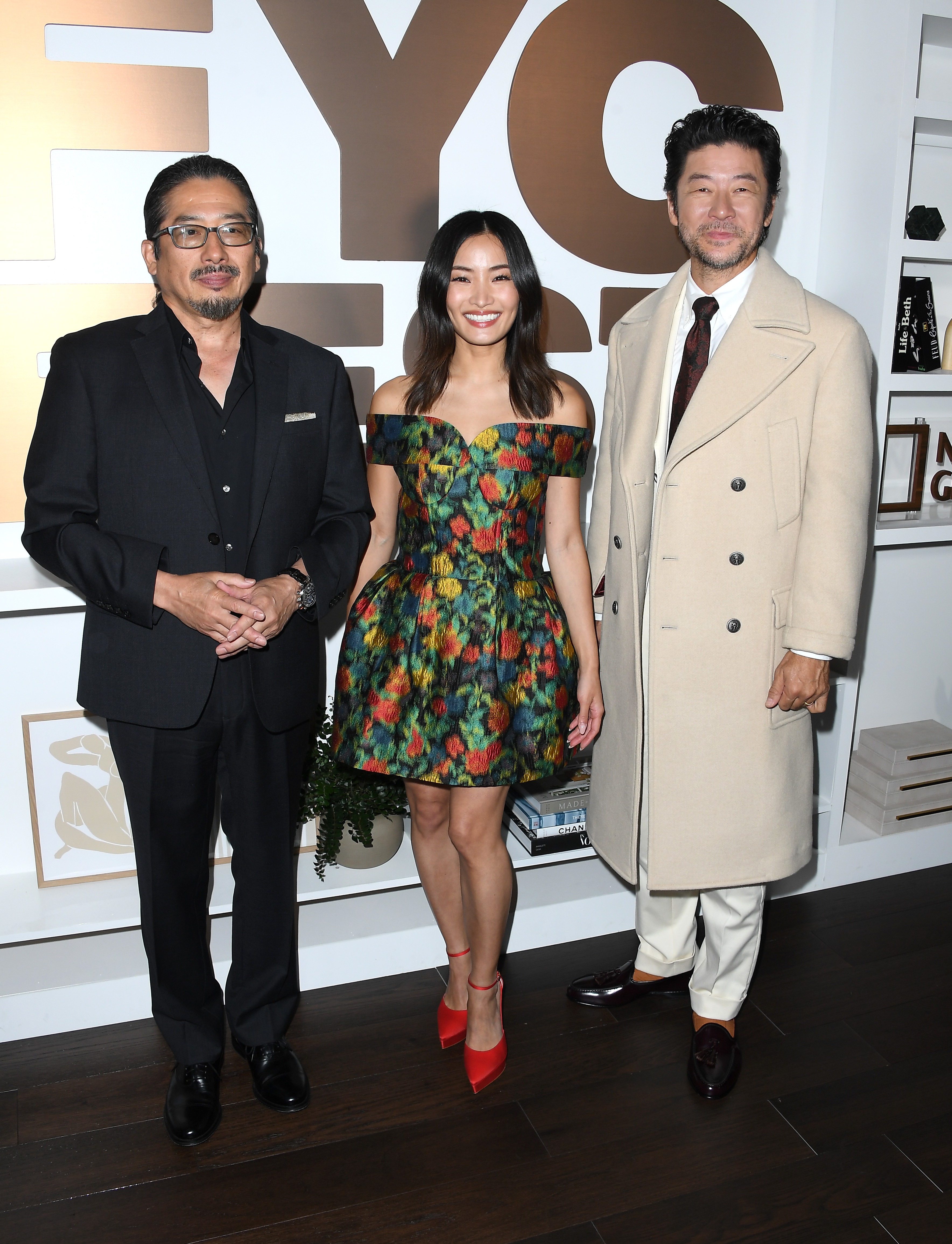 Three people are standing in front of a wall with large text. The woman in the center wears a strapless floral dress with red heels. The men wear dark and light suits