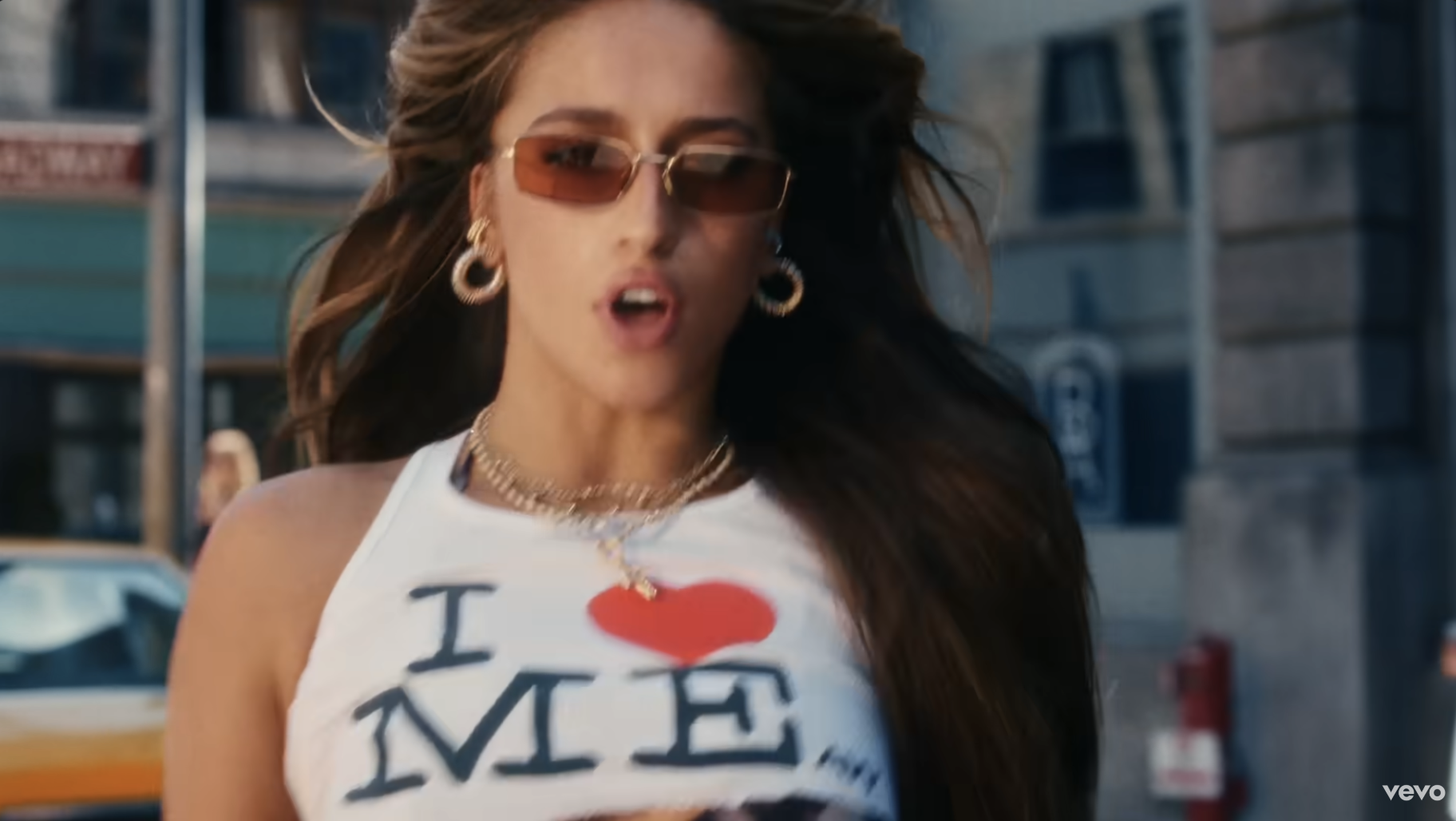 Tate McRae wearing sunglasses, a white tank top with &quot;I ❤️ ME&quot; text, and gold hoop earrings, walks on what appears to be a city street