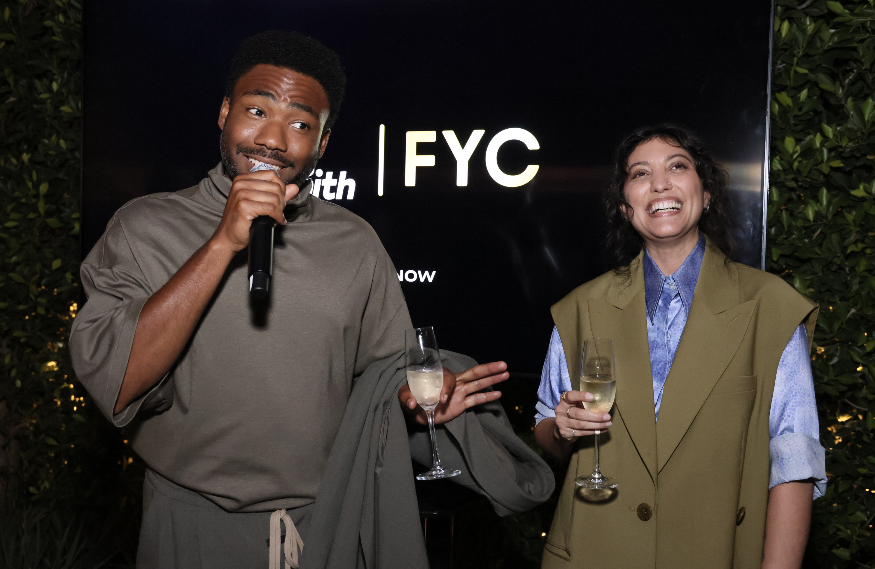 Donald Glover, in a casual robe, and a smiling woman in a green vest and blue shirt, hold champagne glasses at an event with a "FYC" sign in the background