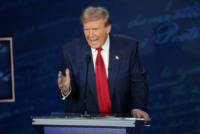 Donald Trump wearing a suit with red tie, speaking at a podium during a public event