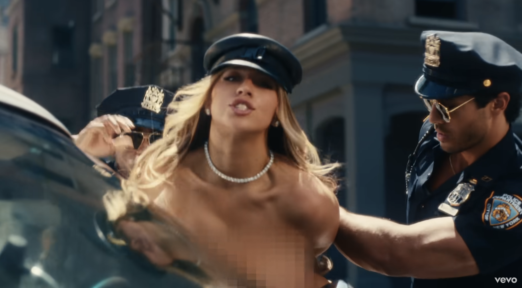 Taylor Swift, in a hat and pearl necklace, is being handcuffed by two police officers on a street