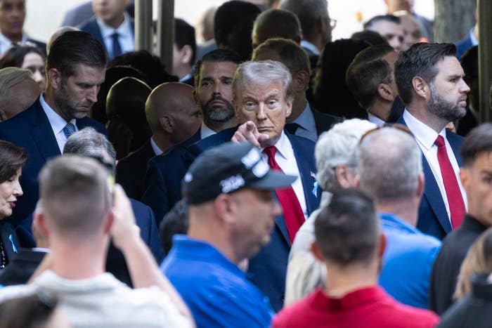 Donald Trump, wearing a suit and red tie, is surrounded by a crowd. Eric Trump and Donald Trump Jr. are also present in the crowd
