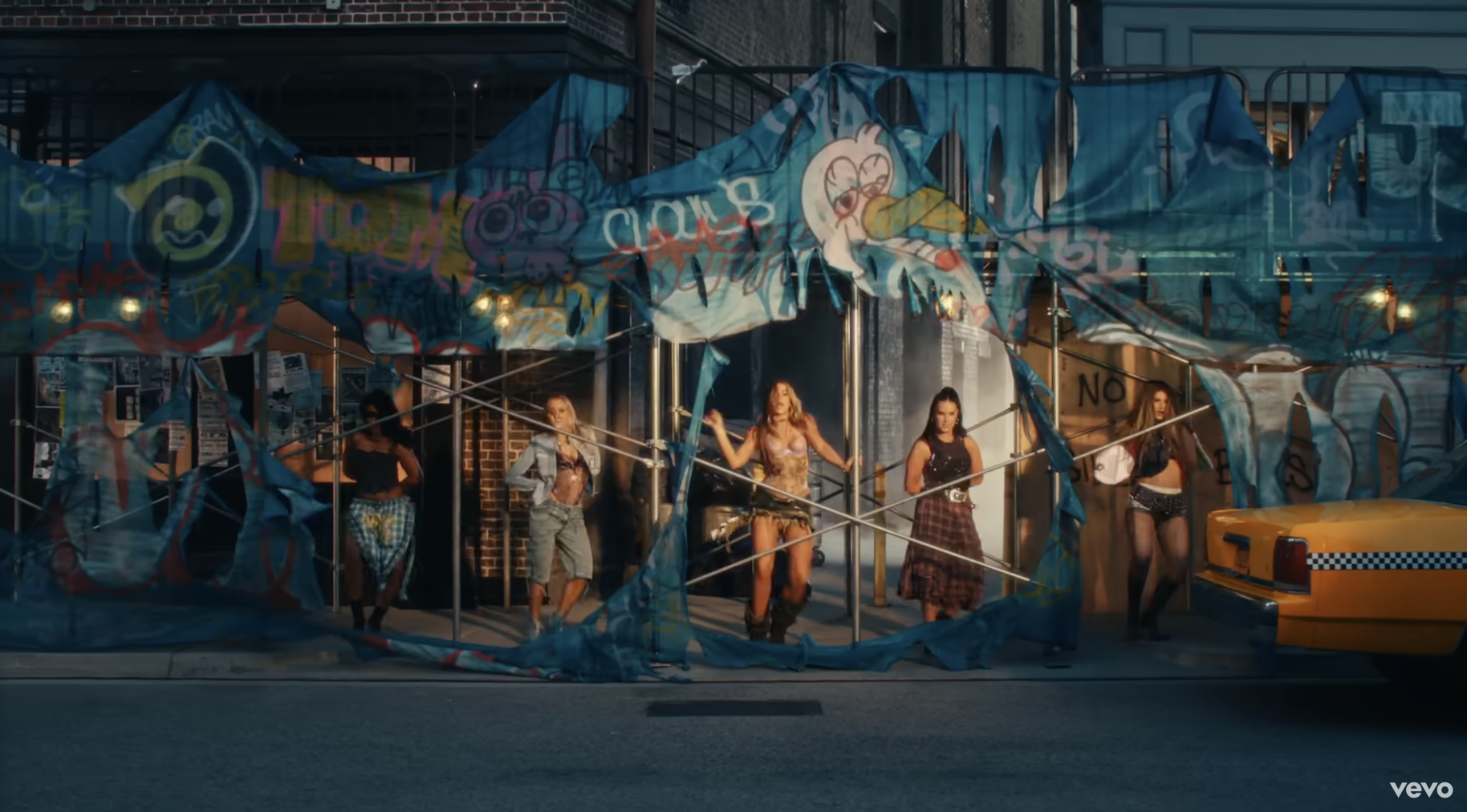 Music video scene featuring several women, including Iggy Azalea, in an urban setting with graffiti, holding poles. A yellow taxi is parked to the right