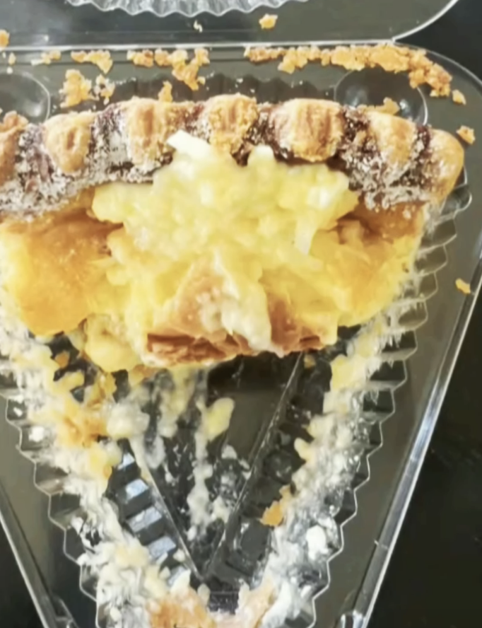 A slice of half-eaten pie with a flaky crust and creamy filling in a plastic container on a dark surface