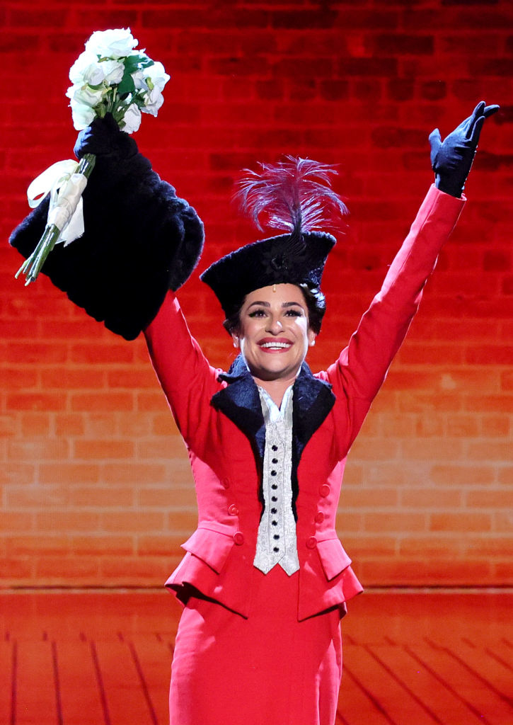 Lea Michele in a stage costume with a hat and gloves, holding a bouquet of flowers, performing a celebratory pose on stage