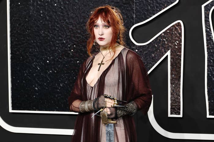 Chappell Roan stands on the red carpet in a stylish sheer outfit with gloves, accessorized with a cross necklace