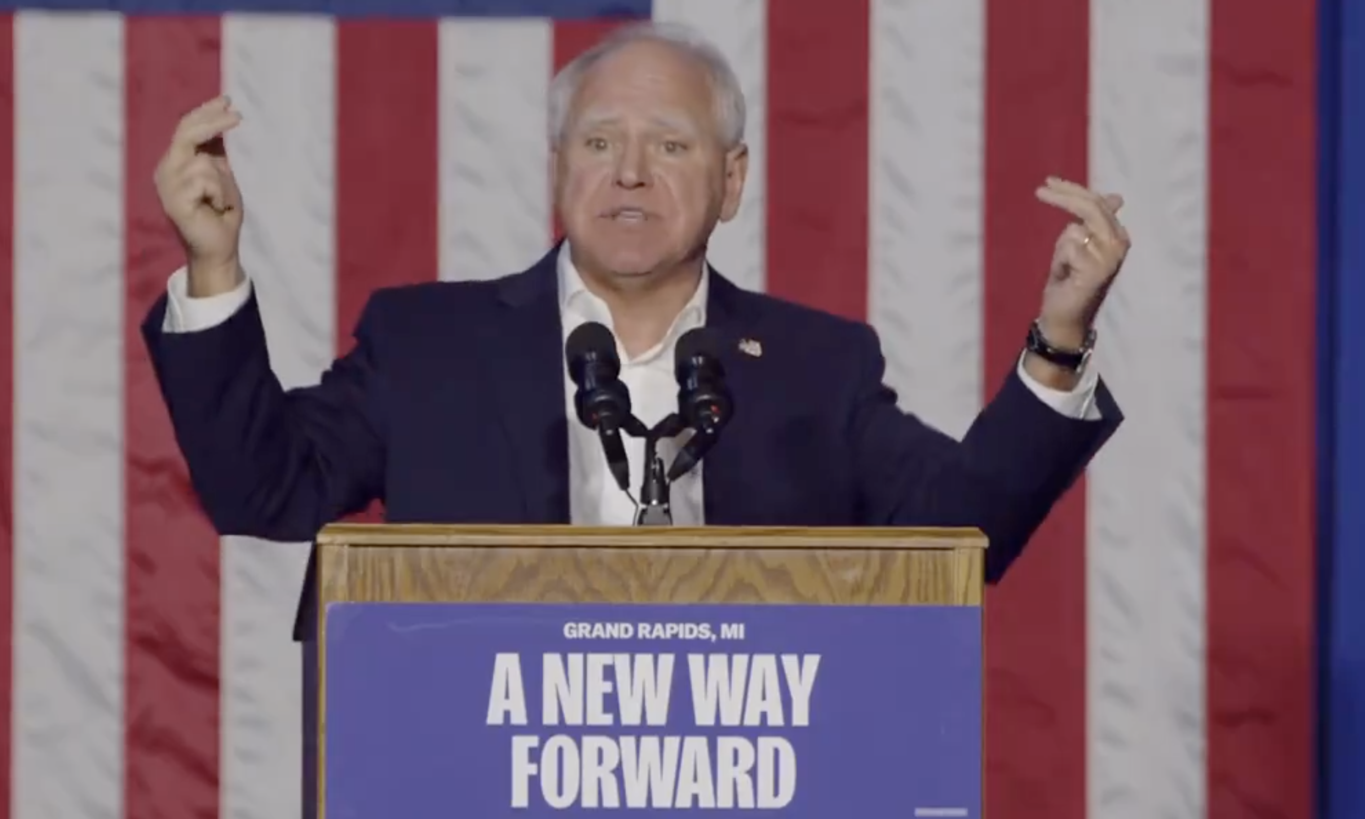 A man is speaking passionately at a podium with a sign that reads "Grand Rapids, MI A New Way Forward." An American flag is in the background