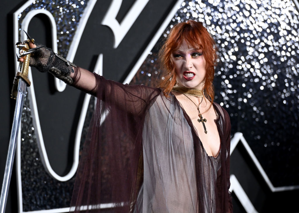 Chappell person on a red carpet, posing with a sword, wearing a sheer, flowy dress and fingerless gloves, with a cross necklace