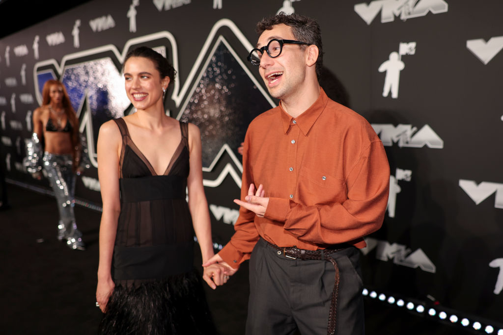 Margaret Qualley and Jack Antonoff holding hands, smiling at each other on the red carpet at the VMAs. Margaret wears a sleek dress, Jack wears a button up shirt and slacks
