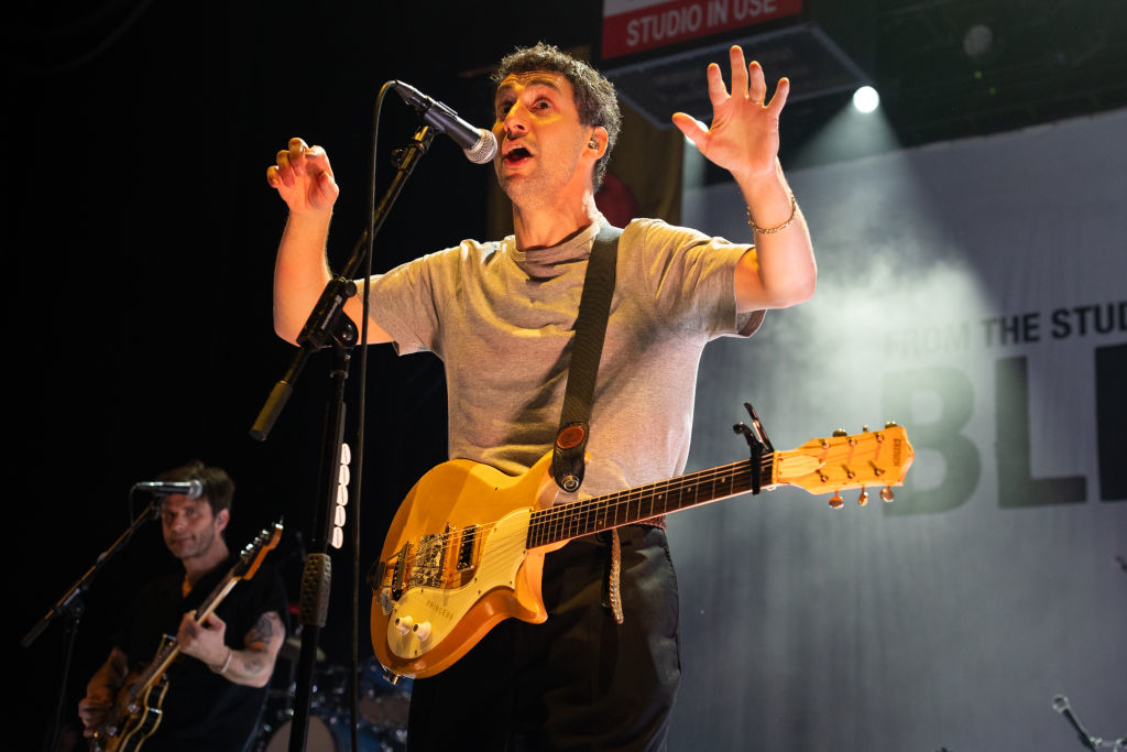 Jack Antonoff performing on stage with a guitar, lead singer of a band. Poster partially visible in the background