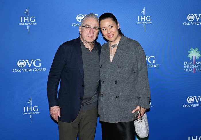 Robert De Niro and Tiffany Chen stand together, smiling at the Palm Springs International Film Festival, with Tiffany holding a clutch