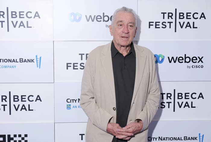 Robert De Niro at the Tribeca Film Festival, wearing a light-colored jacket over a dark shirt, standing in front of a sponsor backdrop
