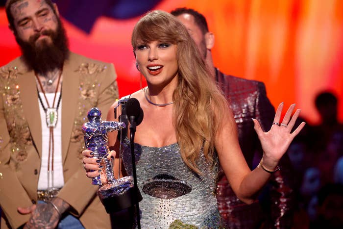 Taylor Swift holds a trophy and speaks at a microphone with Post Malone and another person standing behind her at an event