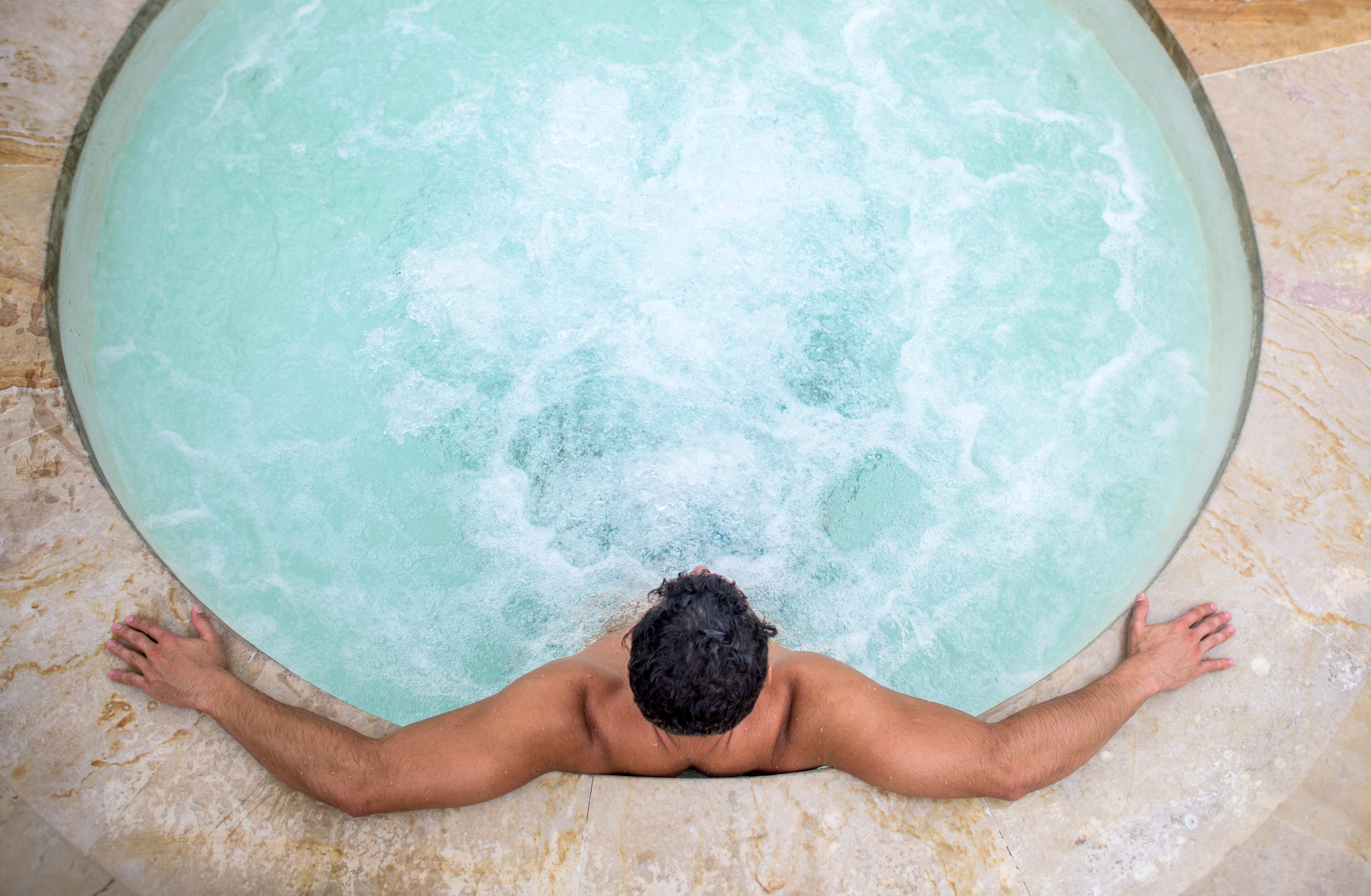 A person is relaxing in a hot tub with their arms stretched out on the edge. Their back is turned to the camera, so their face is not visible