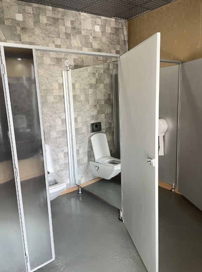 A clean public restroom with stalls. The door to one stall is open, revealing a toilet. The restroom has a modern design with tiled walls and partitioned areas