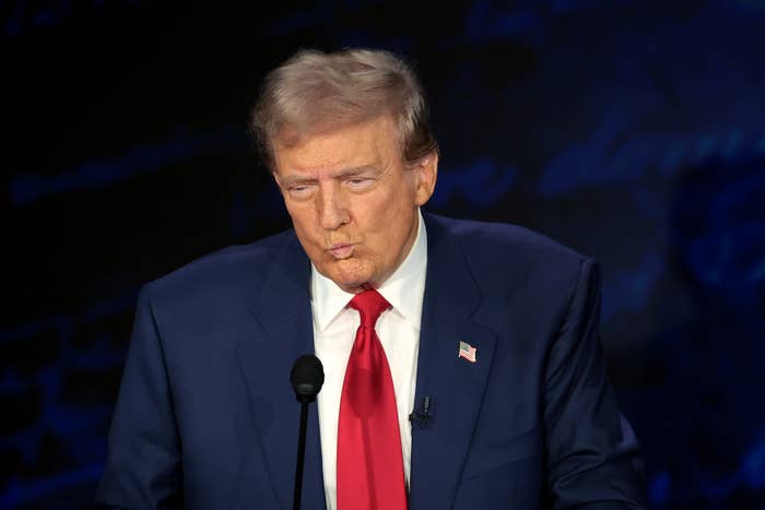Donald Trump, wearing a suit, red tie, and a lapel pin, is making a pursed lip expression during a speaking event