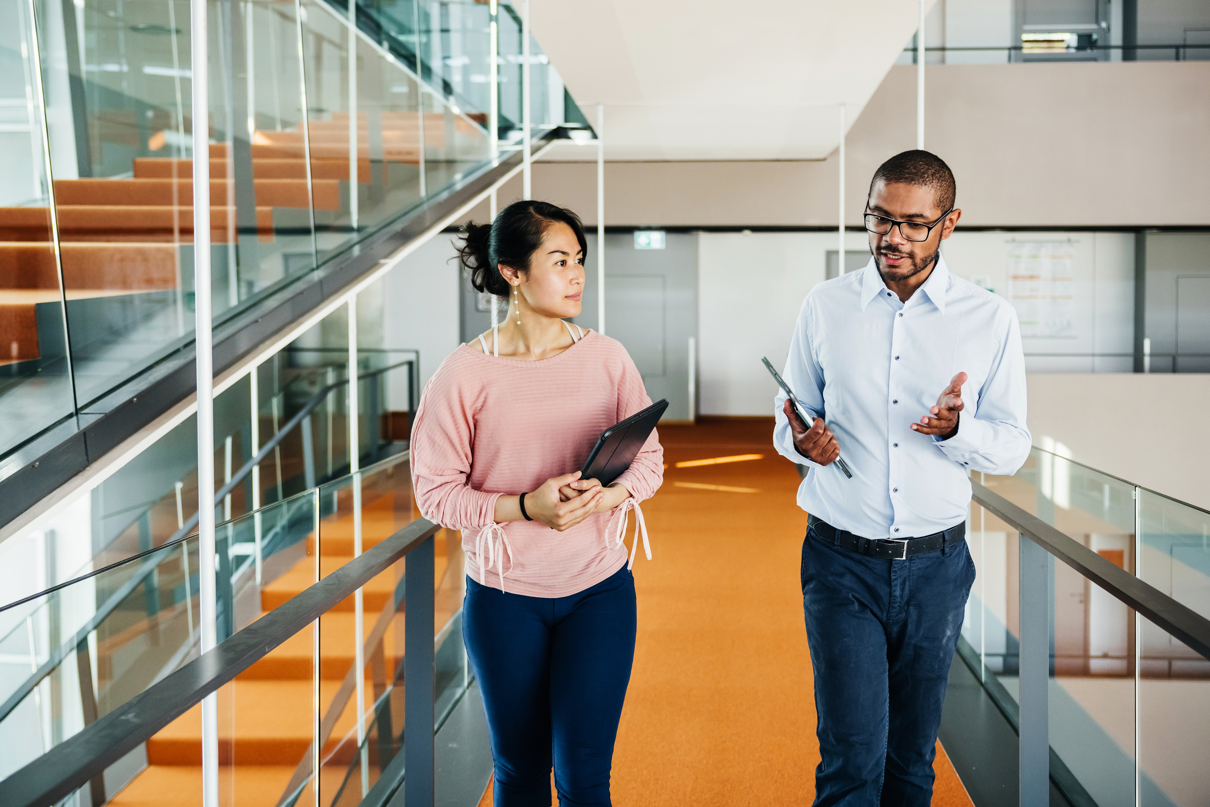 Two professionals engage in conversation while walking down an office corridor. One holds a tablet, the other a folder. Both are casually dressed