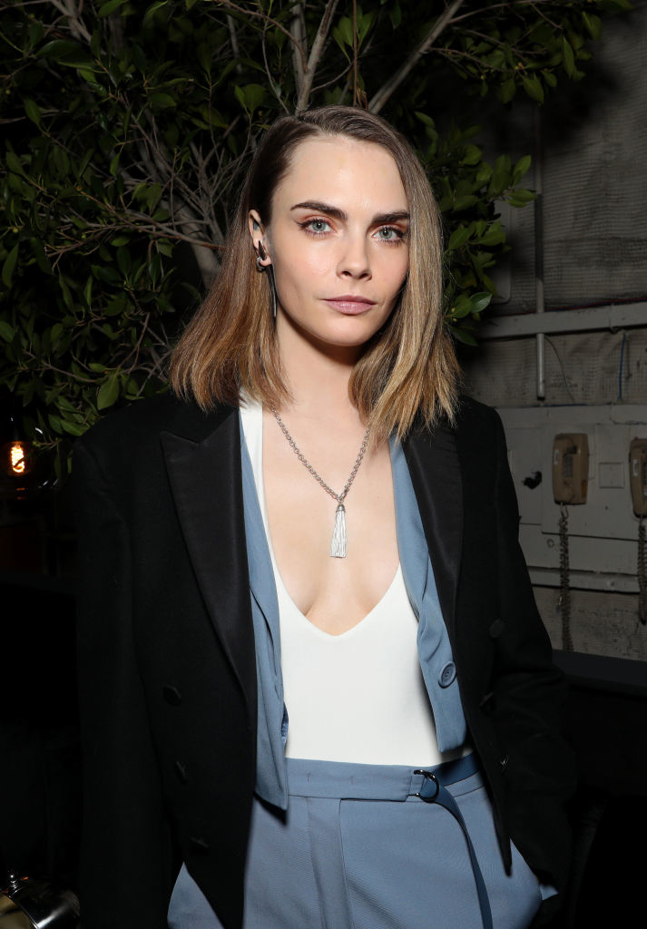 Cara Delevingne in a black blazer over a light-colored deep V-neck top and blue trousers, with a silver necklace, standing indoors in front of a tree