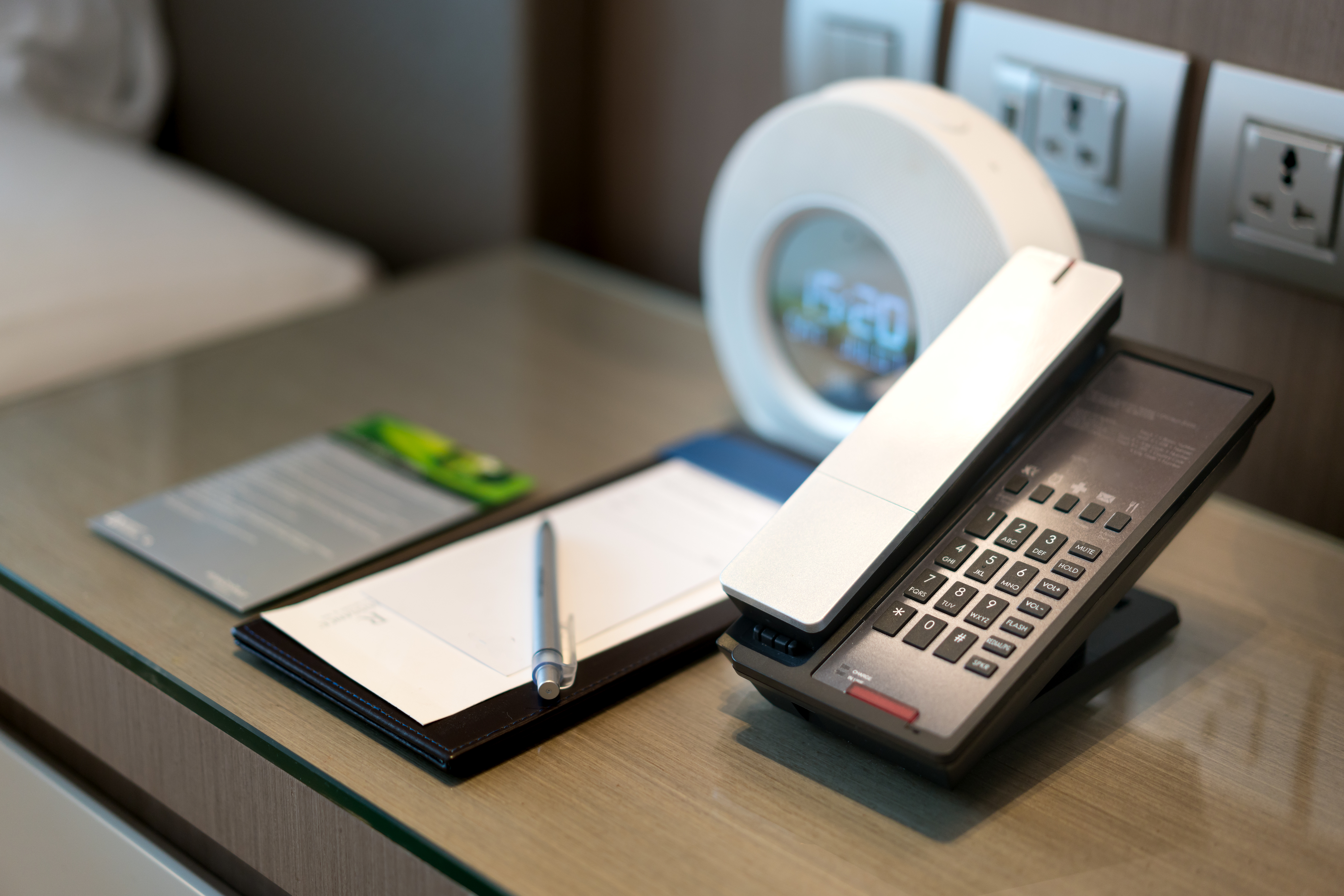 A hotel room's bedside table with a telephone, notepad, pen, and digital clock