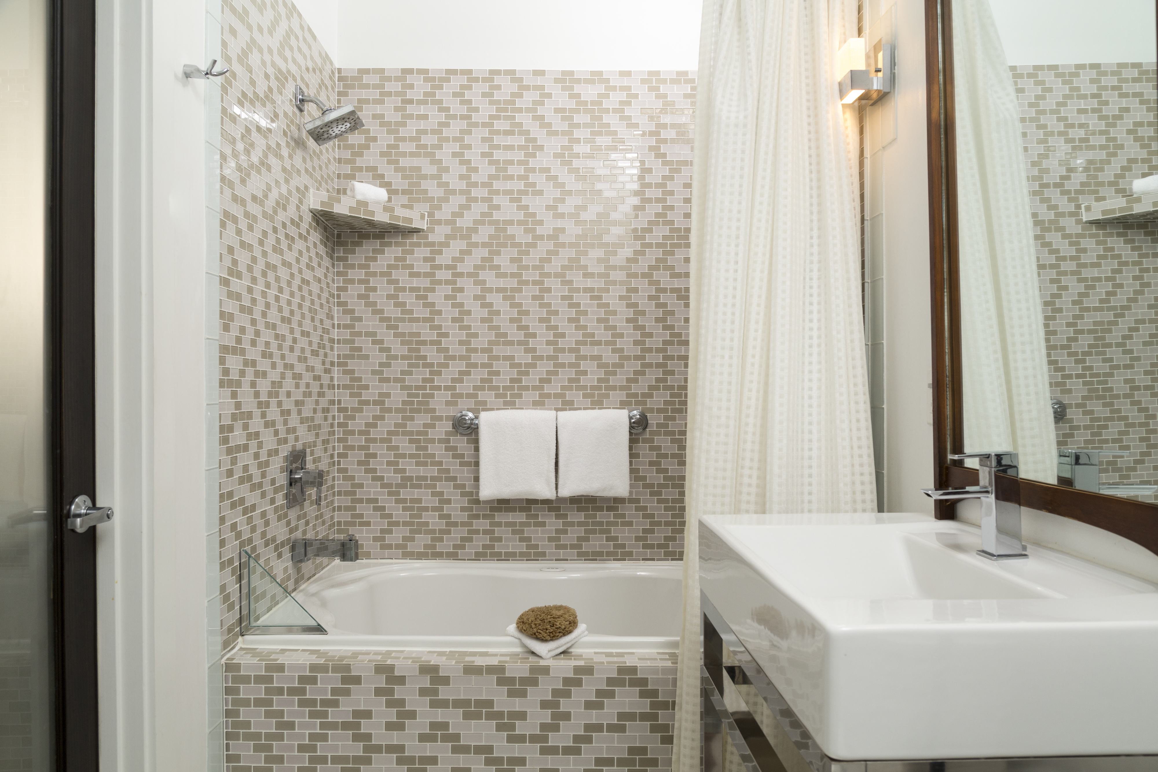 Modern bathroom with mosaic tile walls featuring a bathtub, wall-mounted shelves, a rainfall showerhead, a white sink with a silver faucet, and a wall mirror