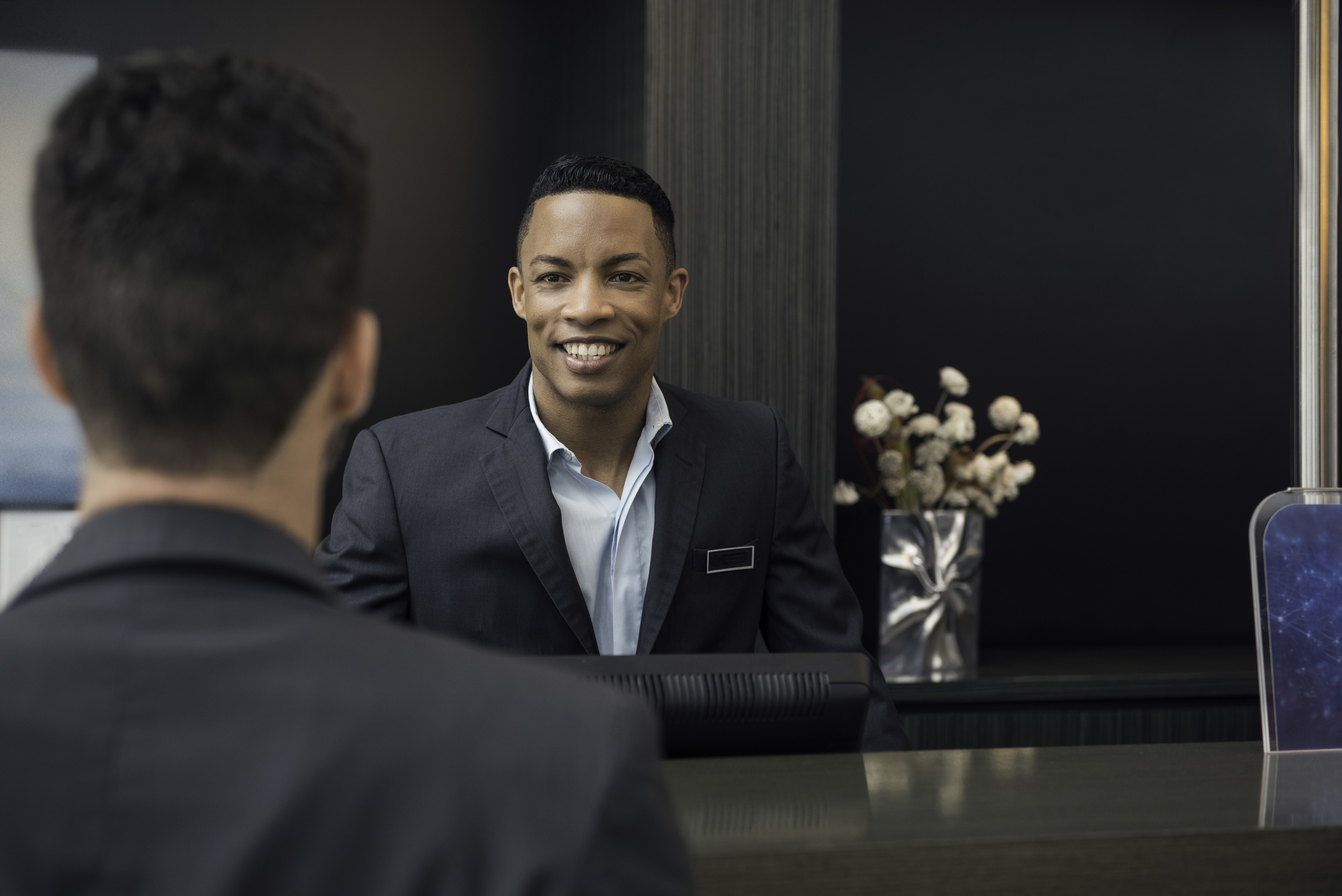A hotel receptionist with a name tag assists a guest at the front desk. The receptionist is smiling and wearing a suit
