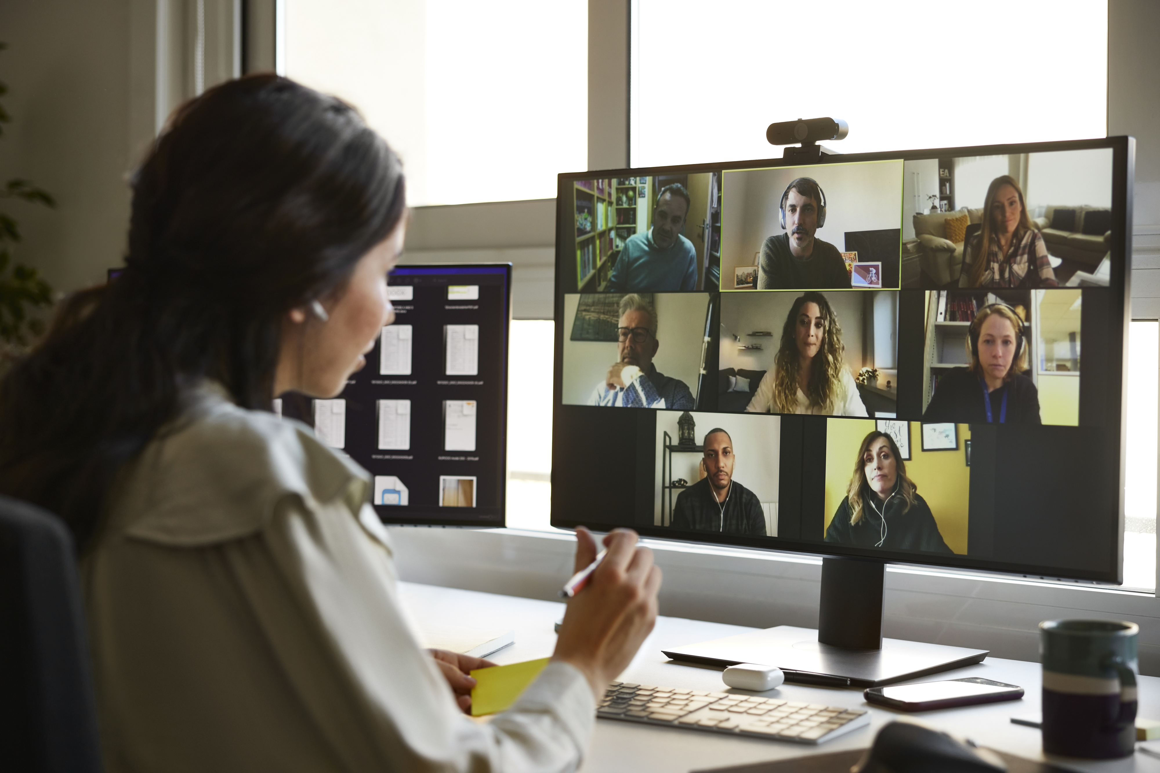 Person in a home office participates in a virtual meeting with nine people on a computer screen. Documents and a coffee cup are on the desk