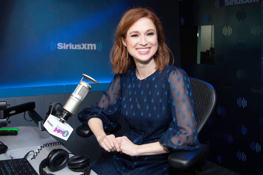 Ellie Kemper sits in a radio studio wearing a stylish, sheer-sleeved dress with a geometric pattern, smiling at the camera in front of a SiriusXM microphone