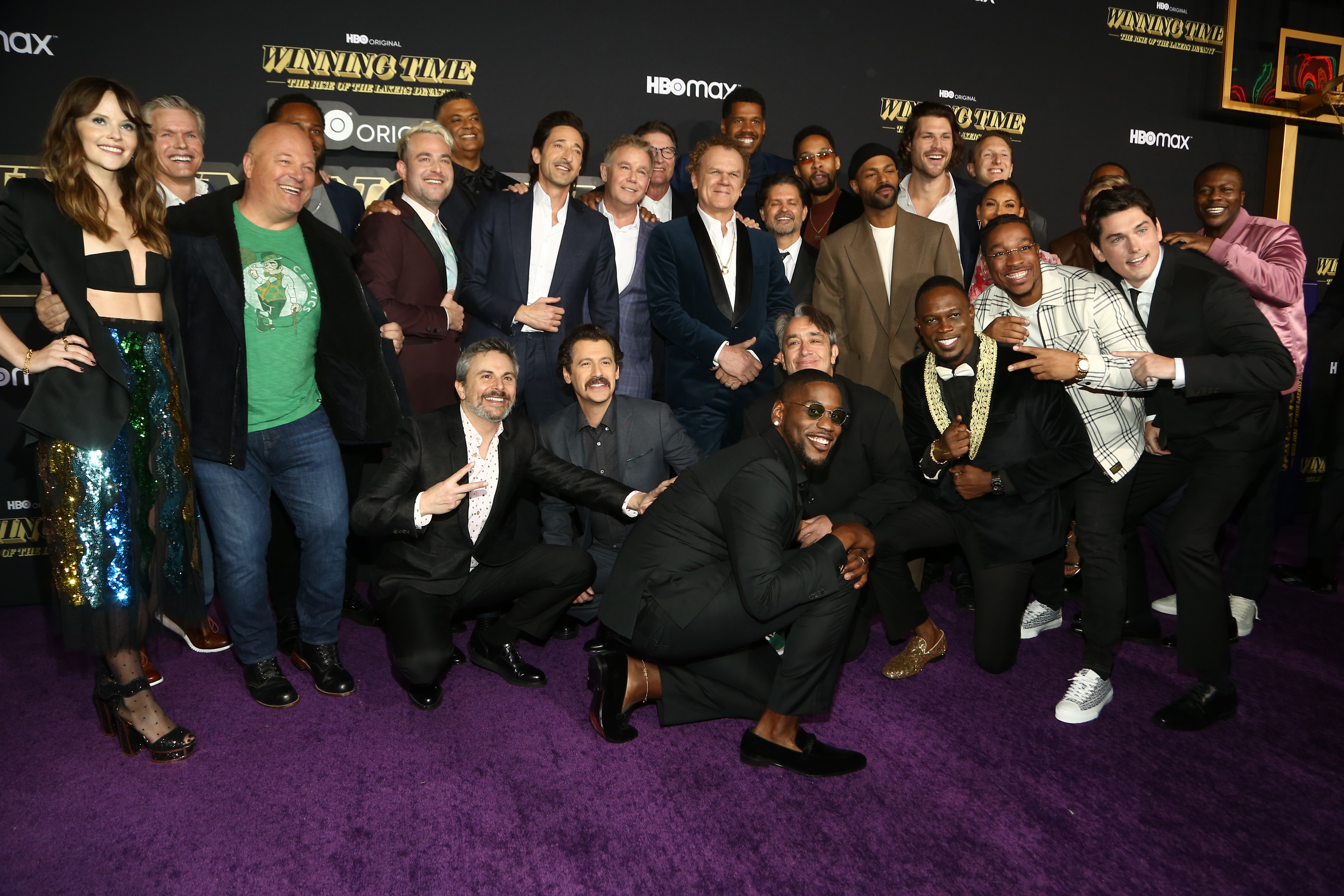 Group photo of cast and crew from "Winning Time: The Rise of the Lakers Dynasty" on red carpet, wearing formal attire including suits and dresses