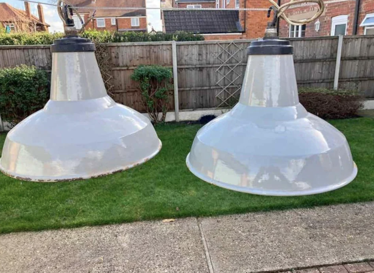 Two large industrial-style pendant light fixtures hanging outdoors in a garden area with grass, a paved path, and a wooden fence in the background