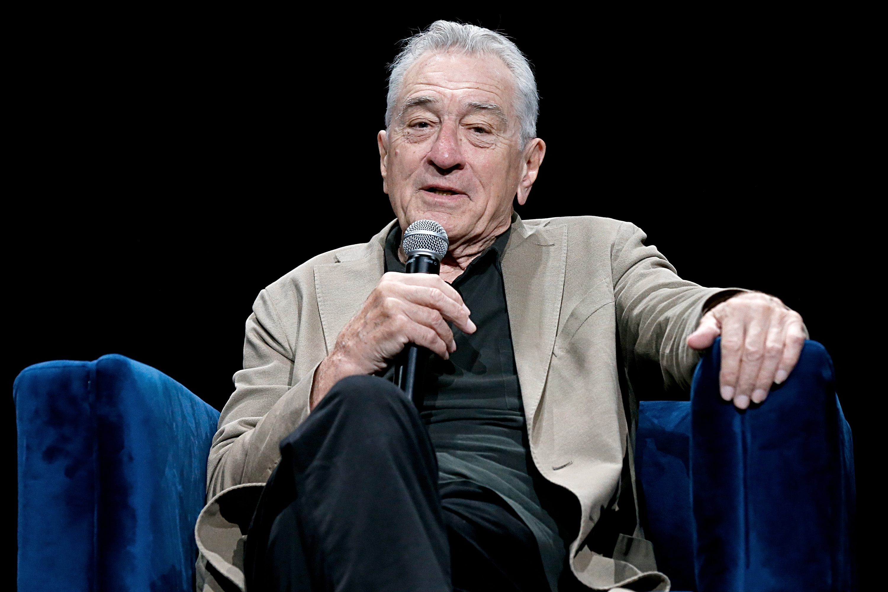 Robert De Niro sits on a chair, holding a microphone, wearing a blazer and dark shirt