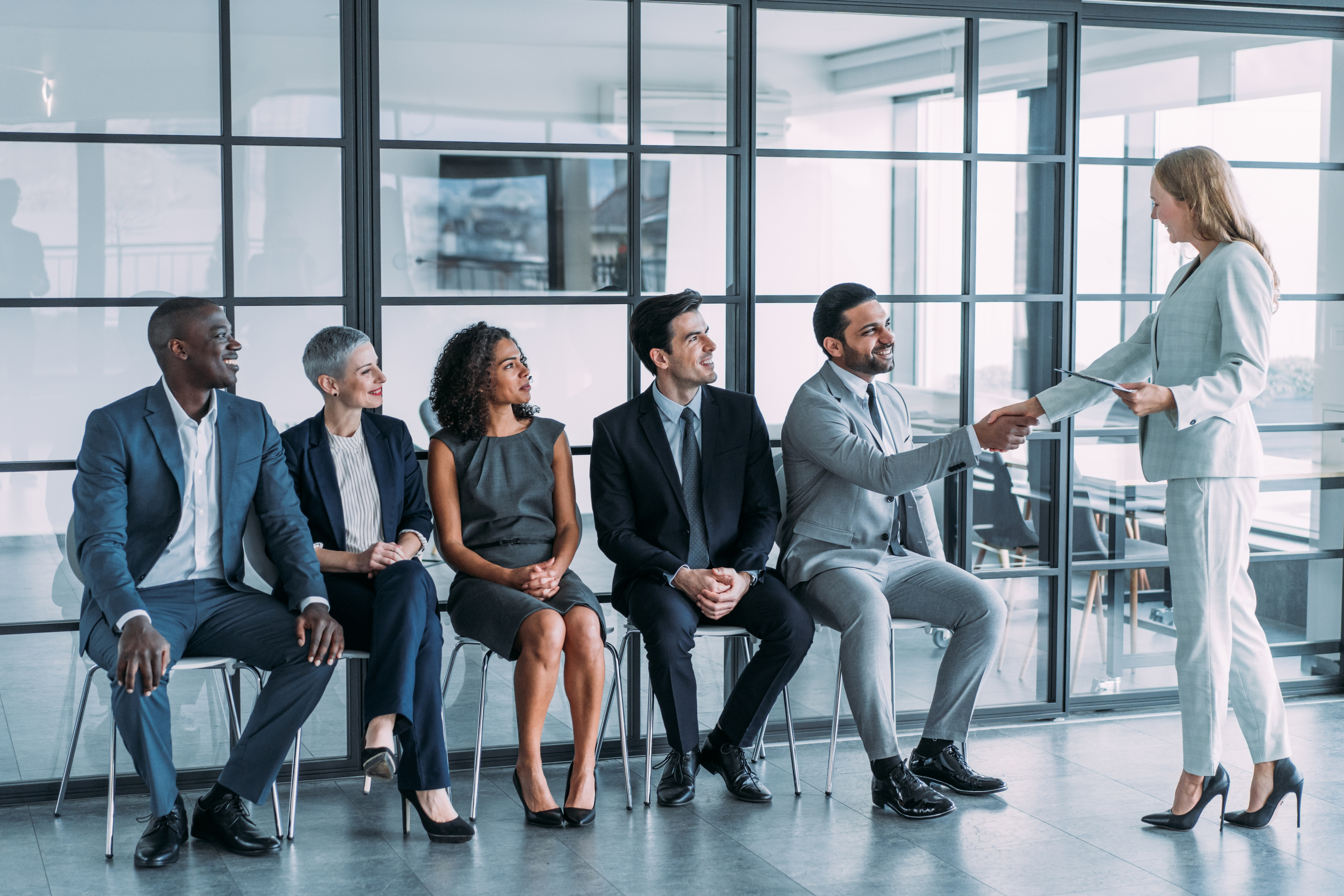 Five professionals sit in a row in an office; the sixth person, standing, hands a document to one seated individual