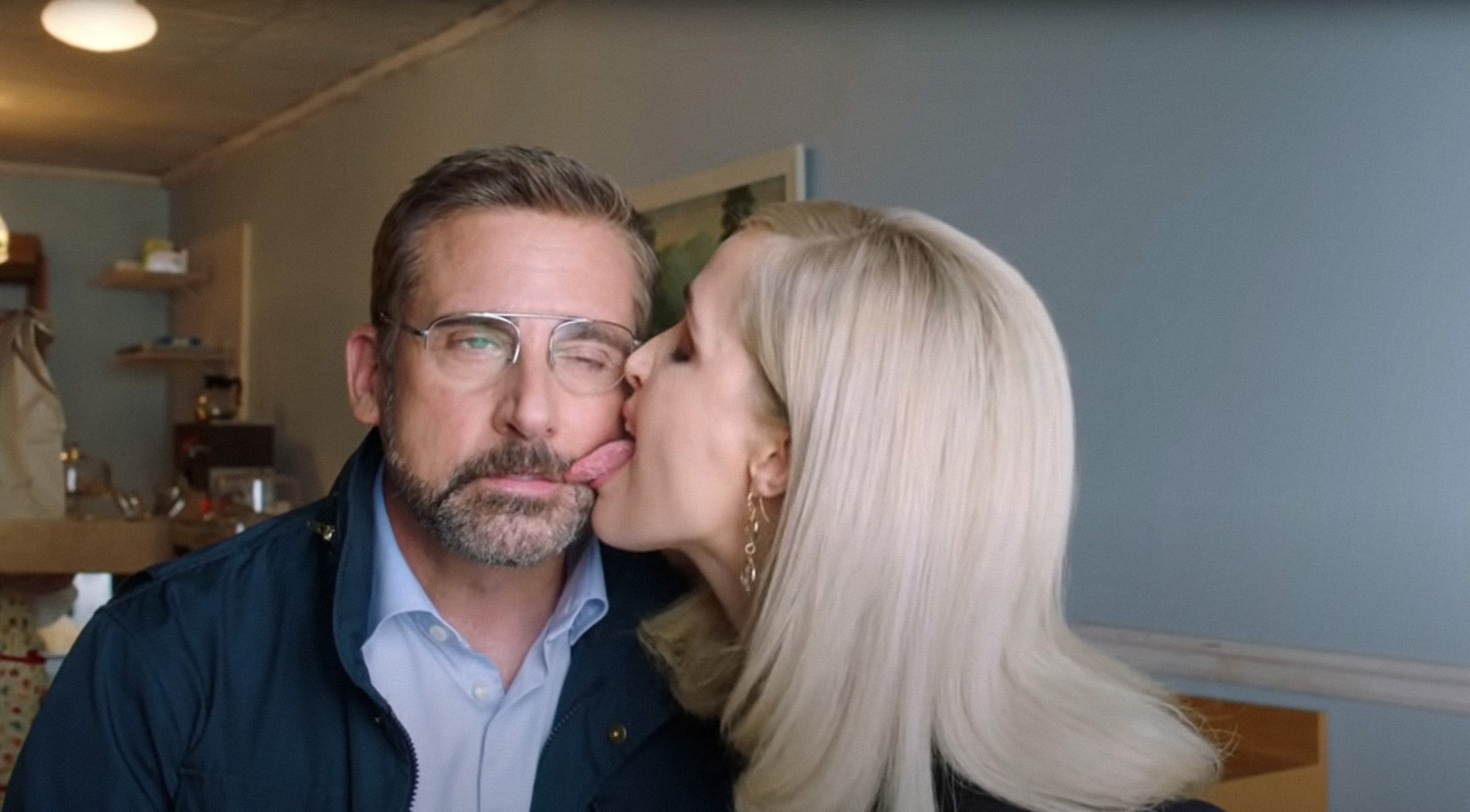 Steve Carell gets his face humorously licked by a woman with long blonde hair in a casual indoor setting