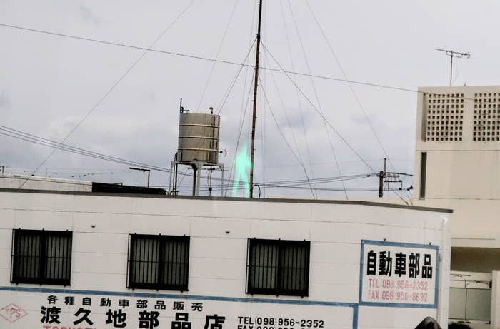 A multi-story building features Japanese signage with phone numbers. Atop the building, a greenish flame-like light appears, seemingly emitting from the roof