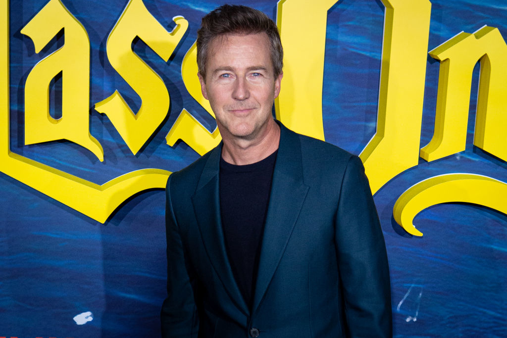 Edward Norton in a dark blazer and black shirt at the "Glass Onion" event. The backdrop features the movie's title in bold, stylized letters
