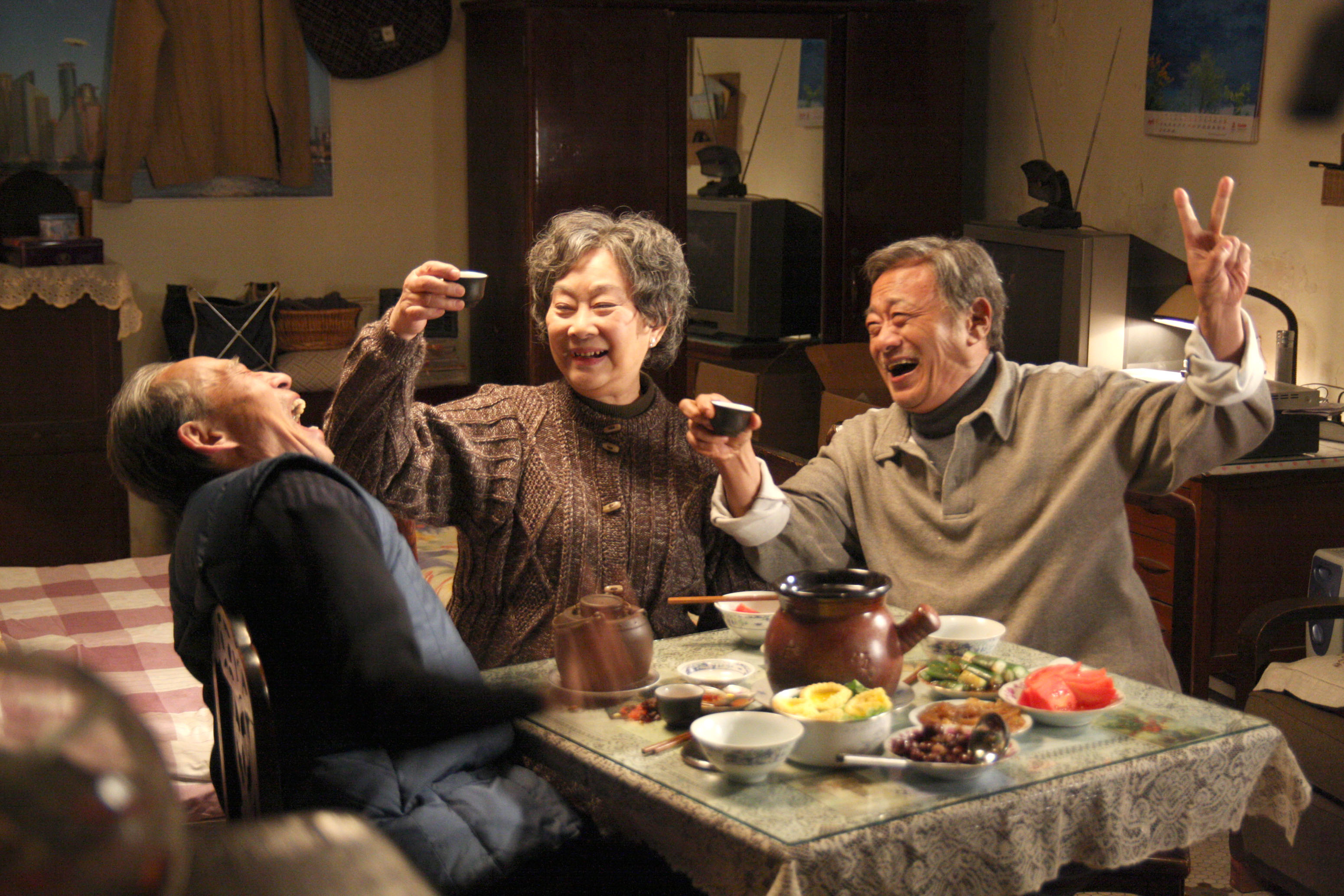Three older adults, all smiling, raise cups in a cheerful toast around a table set with various dishes. The setting appears to be a cozy dining room