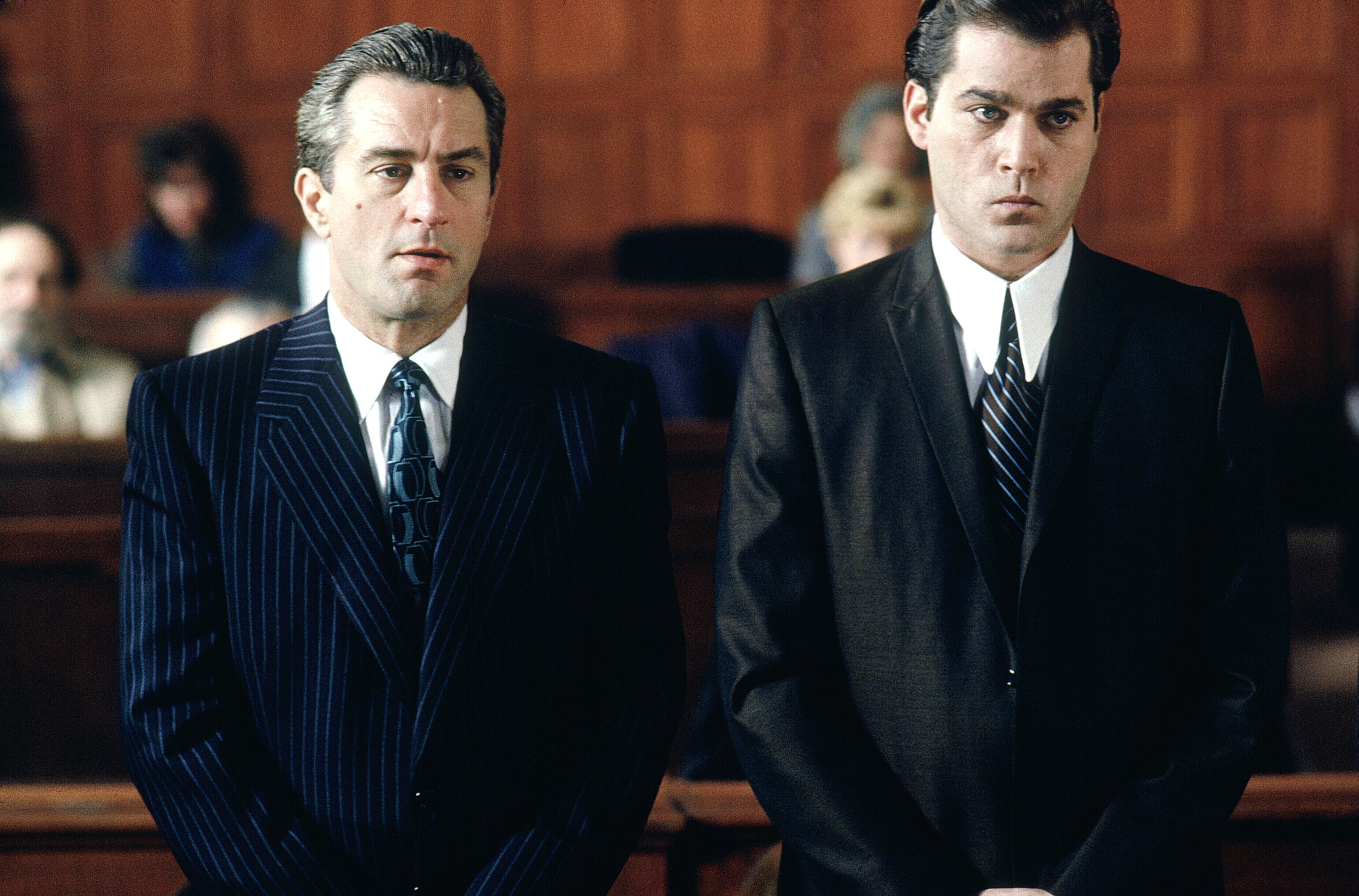 Robert De Niro and Ray Liotta are dressed in suits, standing in a courtroom, appearing serious