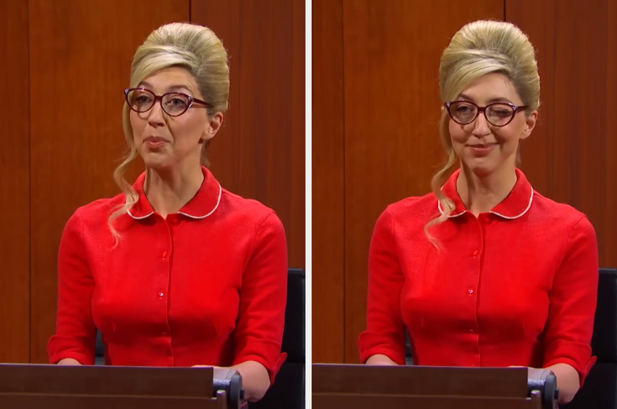 Heidi Gardner wearing glasses and a red dress with a high neckline and buttons sitting at a desk in an SNL skit