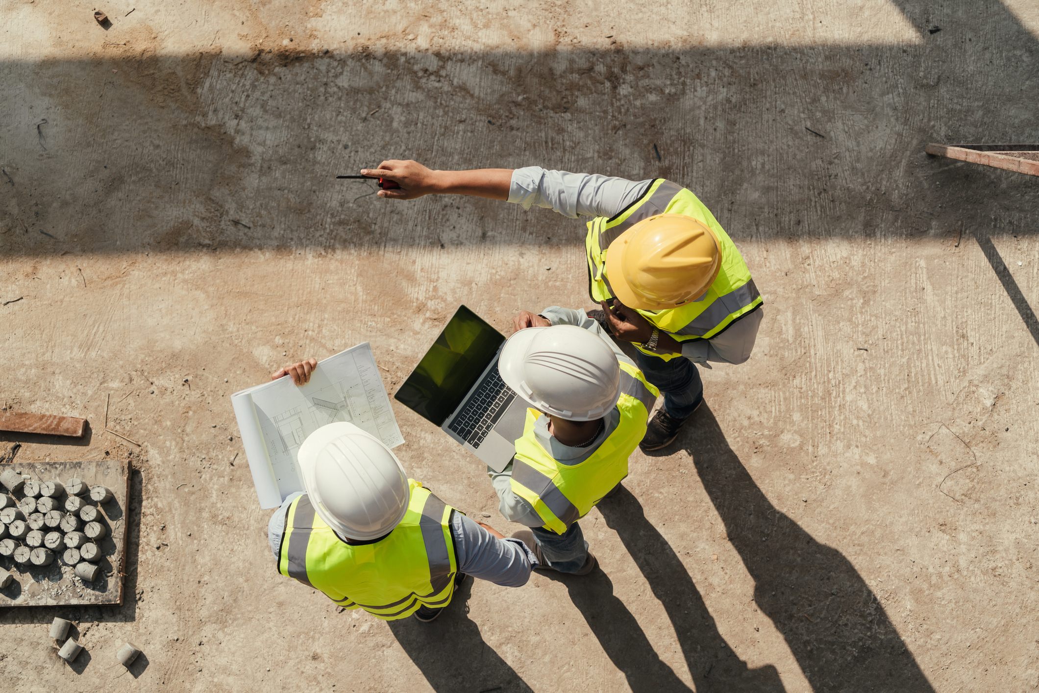Three construction workers in safety vests and helmets review plans and a laptop on-site, with one worker pointing out something in the distance