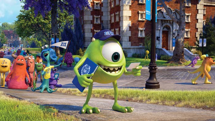 Mike Wazowski in a Monsters University cap walks on campus holding a flag and documents, surrounded by other colorful monster students