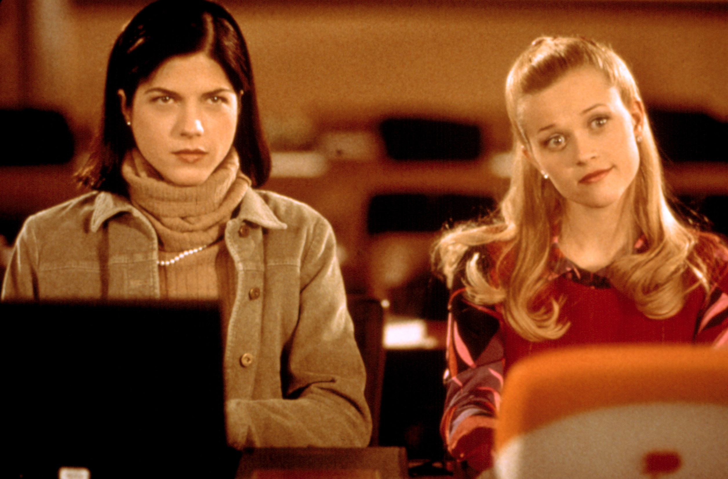 Selma Blair and Reese Witherspoon sit in a classroom, with serious expressions, looking towards the front. Selma wears a turtleneck and cardigan; Reese wears a sweater